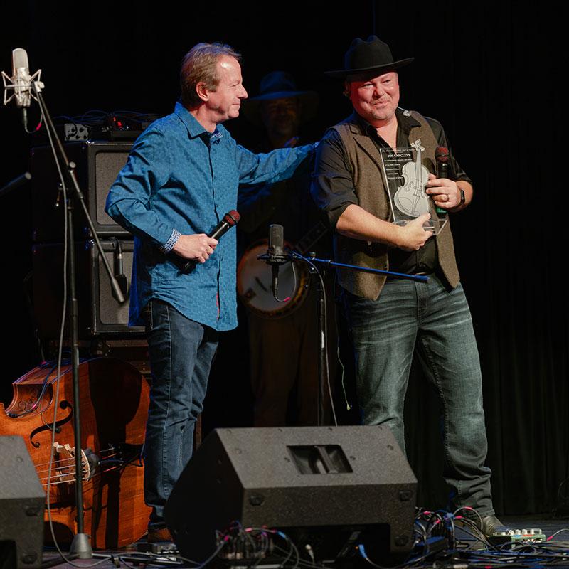 Stuart Duncan (left) congratulates Jim VanCleve after presenting him with his National Fiddler Hall of Fame award at the Appalachian Theatre of the High Country in Boone, North Carolina, on October 4, 2026.  Photo credit: Jamey Booher Photography